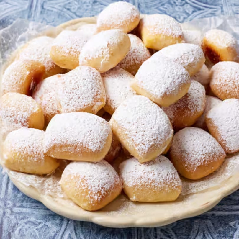A platter of golden-brown beignets, generously dusted with powdered sugar, on a light blue patterned tablecloth.