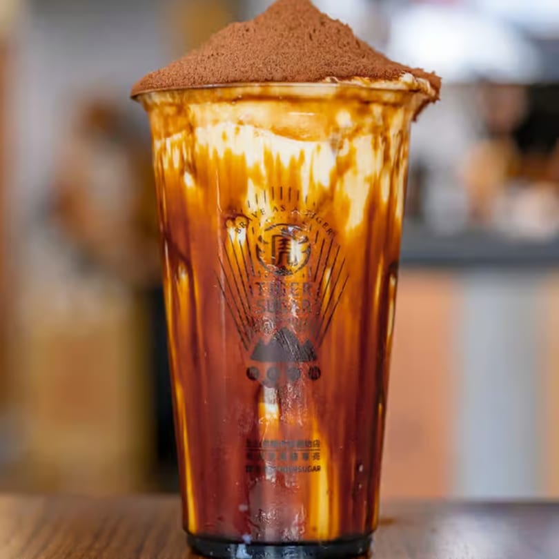Brown Sugar Boba Milk with cream and cocoa powder on top, in a clear plastic cup.
